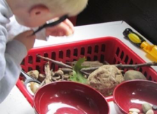 Student peering through a magnifying glass into a basket filled with twigs, leaves, and other natural materials.